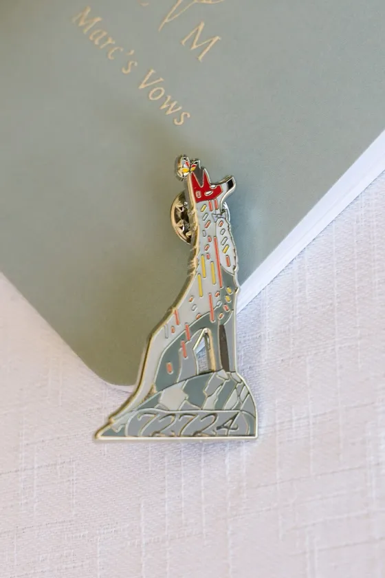 Enamel pin of a howling wolf with a butterfly, engraved 7.27.24, resting on a vow book